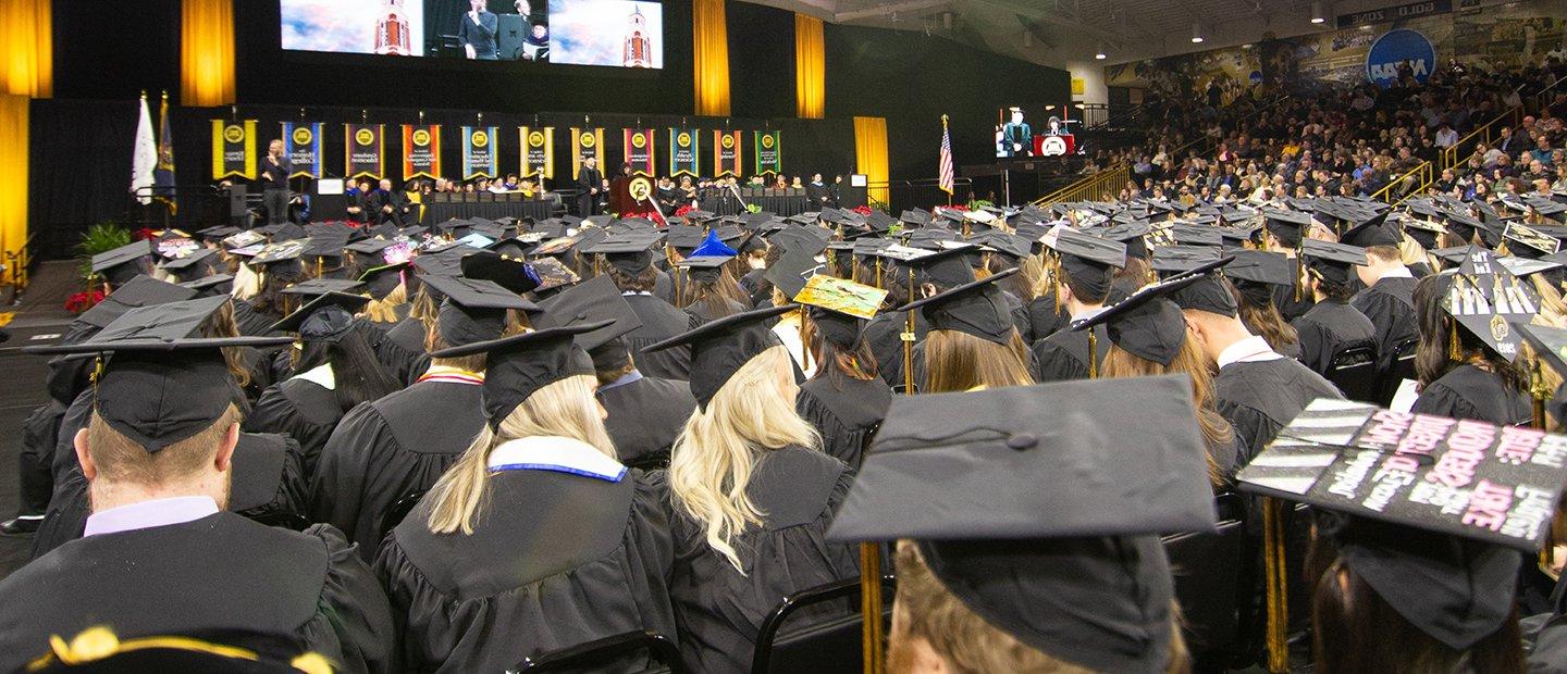 A photo of graduates in caps and gowns, facing a stage.