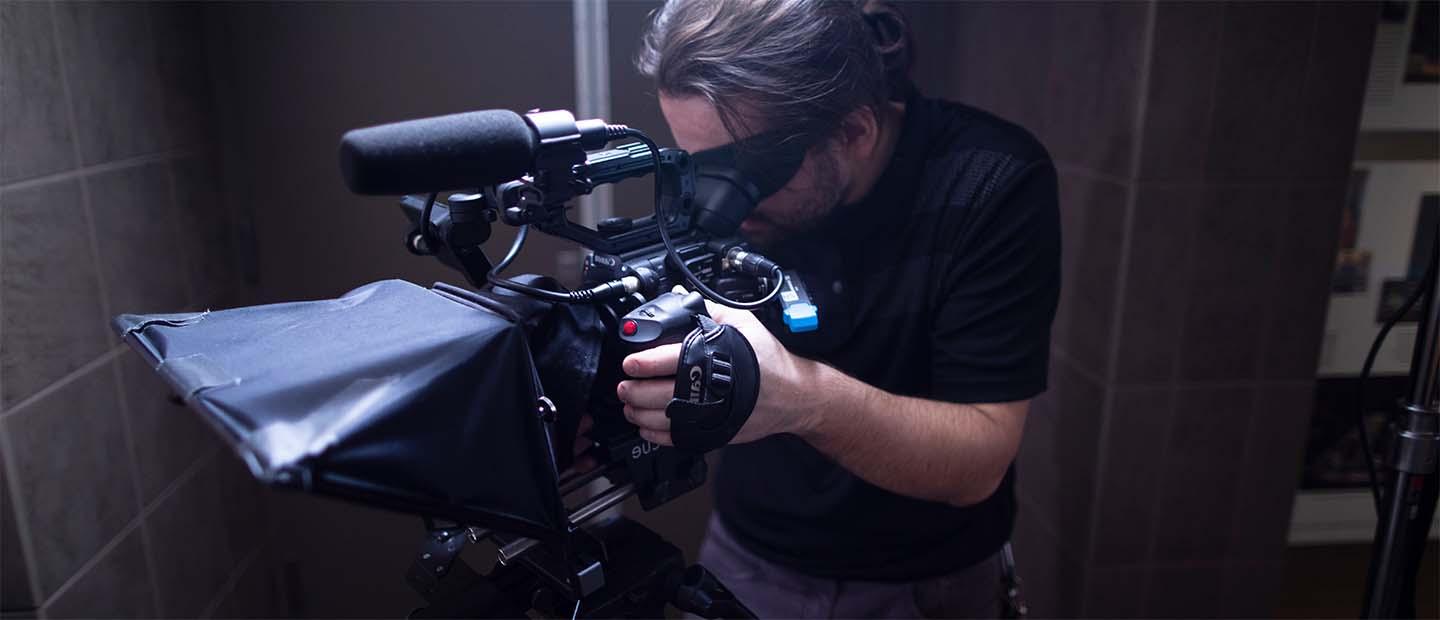  A man standing behind a video camera, filming.