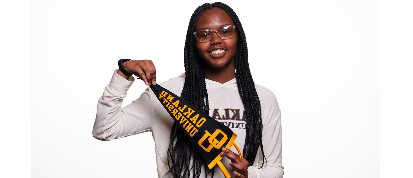 Young woman smiles at the camera. She is wearing and Oakland University shirt and is holding an O U pennant.